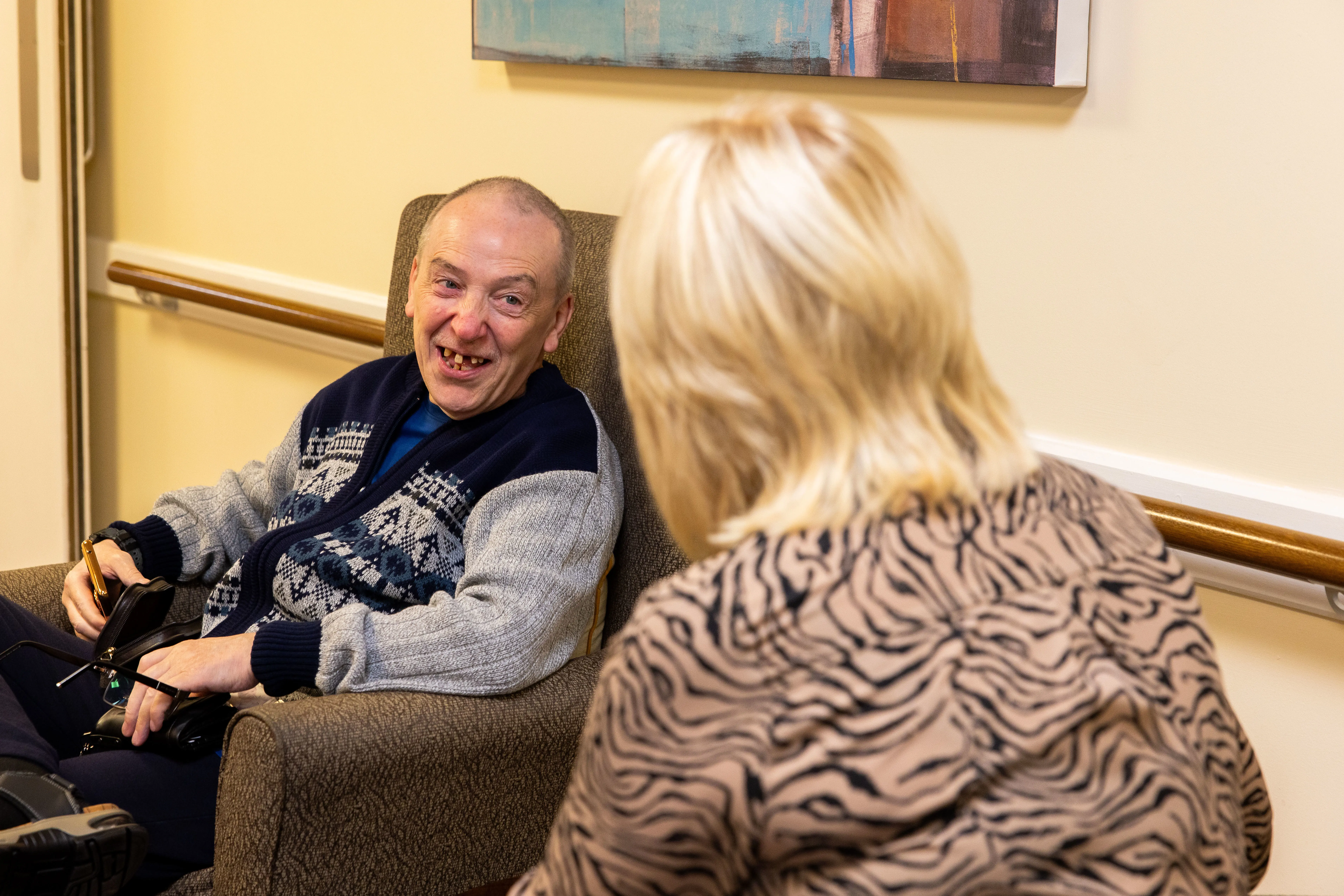 Older man sat down in an armchair, speaking to a staff member laughing together. 