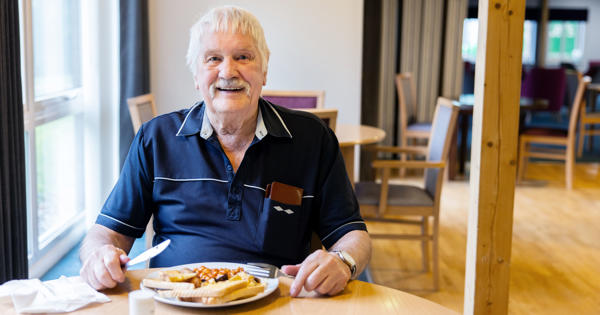 Older man sat at a dining table, with a breakfast on a plate in front of him, looking at the camera and smiling. 